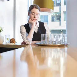 Fed up waitress at work. She has her eyes closed and is resting her head on her hand at the bar, with a tray of dirty glasses in front of her.