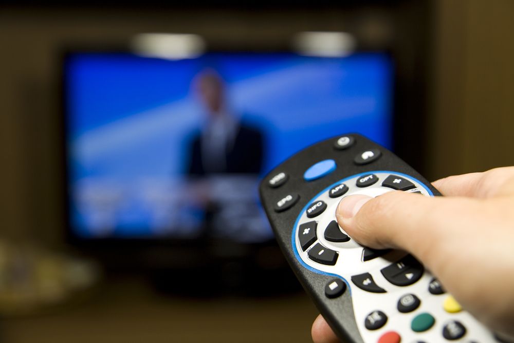 Hand holding TV remote control with a television in the background. Close up.