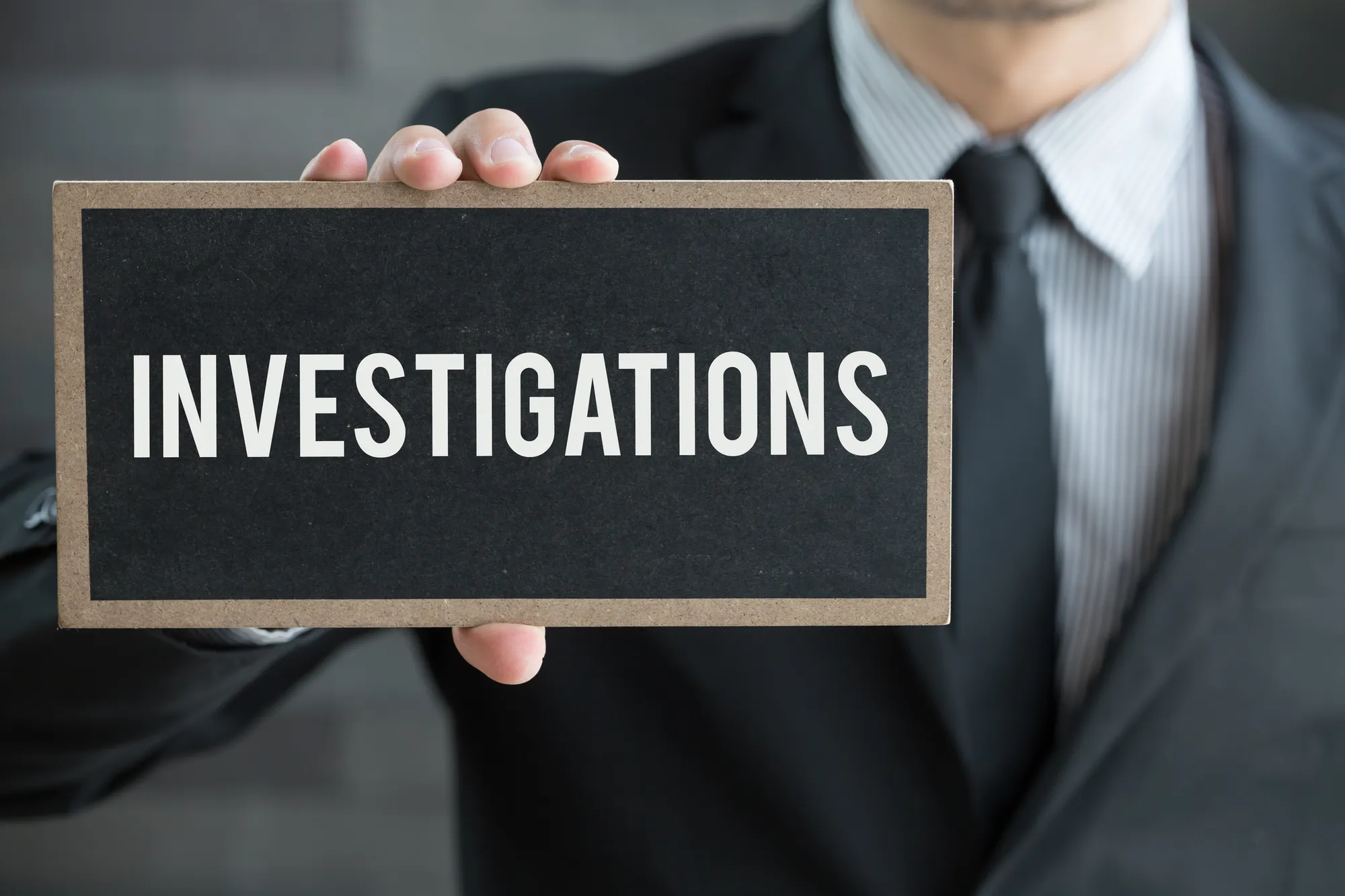 A man in suit holding out an "Investigations" sign in one hand — lawsuit investigations, class action investigation