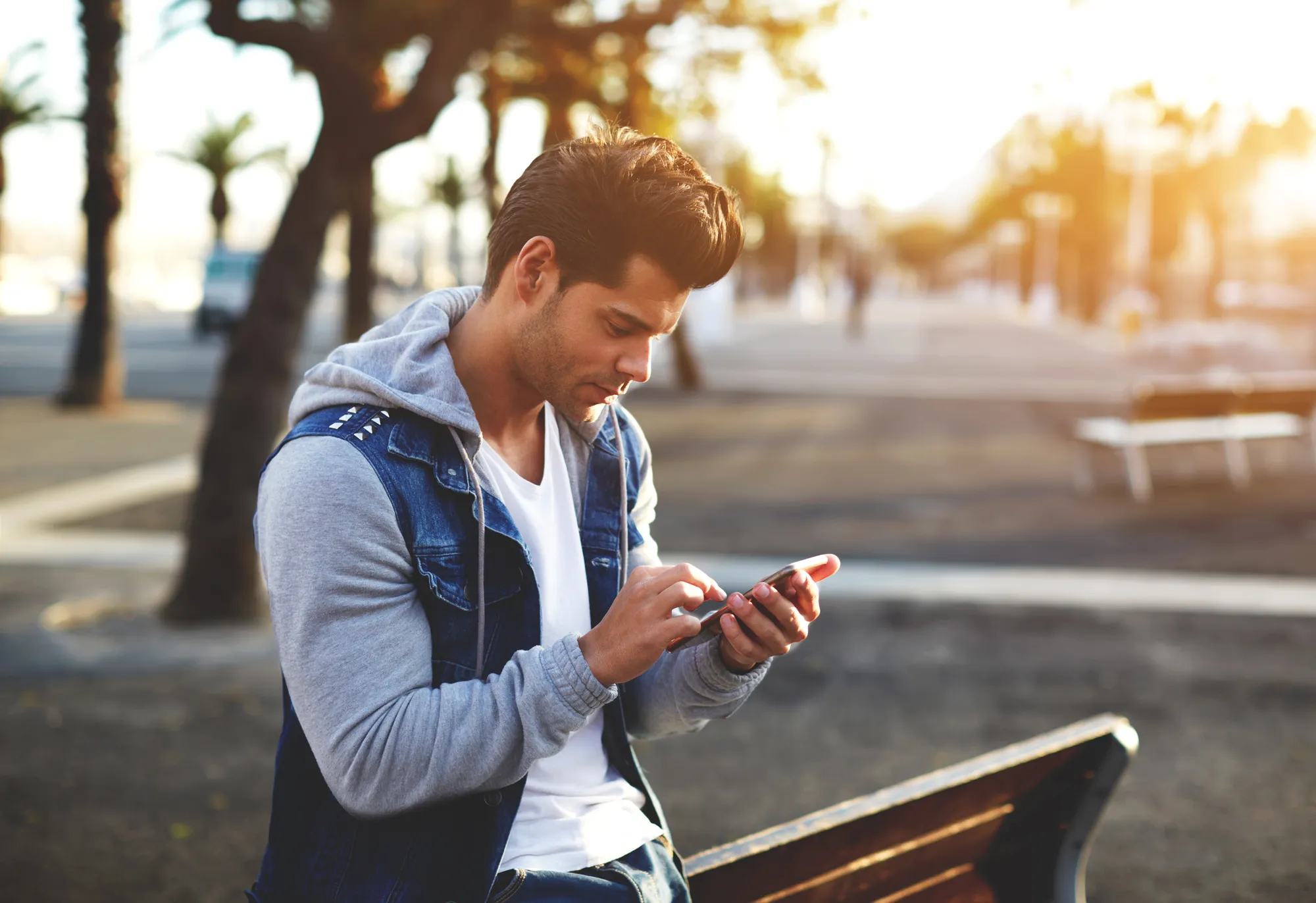 Portrait of handsome young man sending a text message while standing outside, male student reading text messages outdoors, stylish brunette hipster using cell phone at sunny evening, flare sunshine