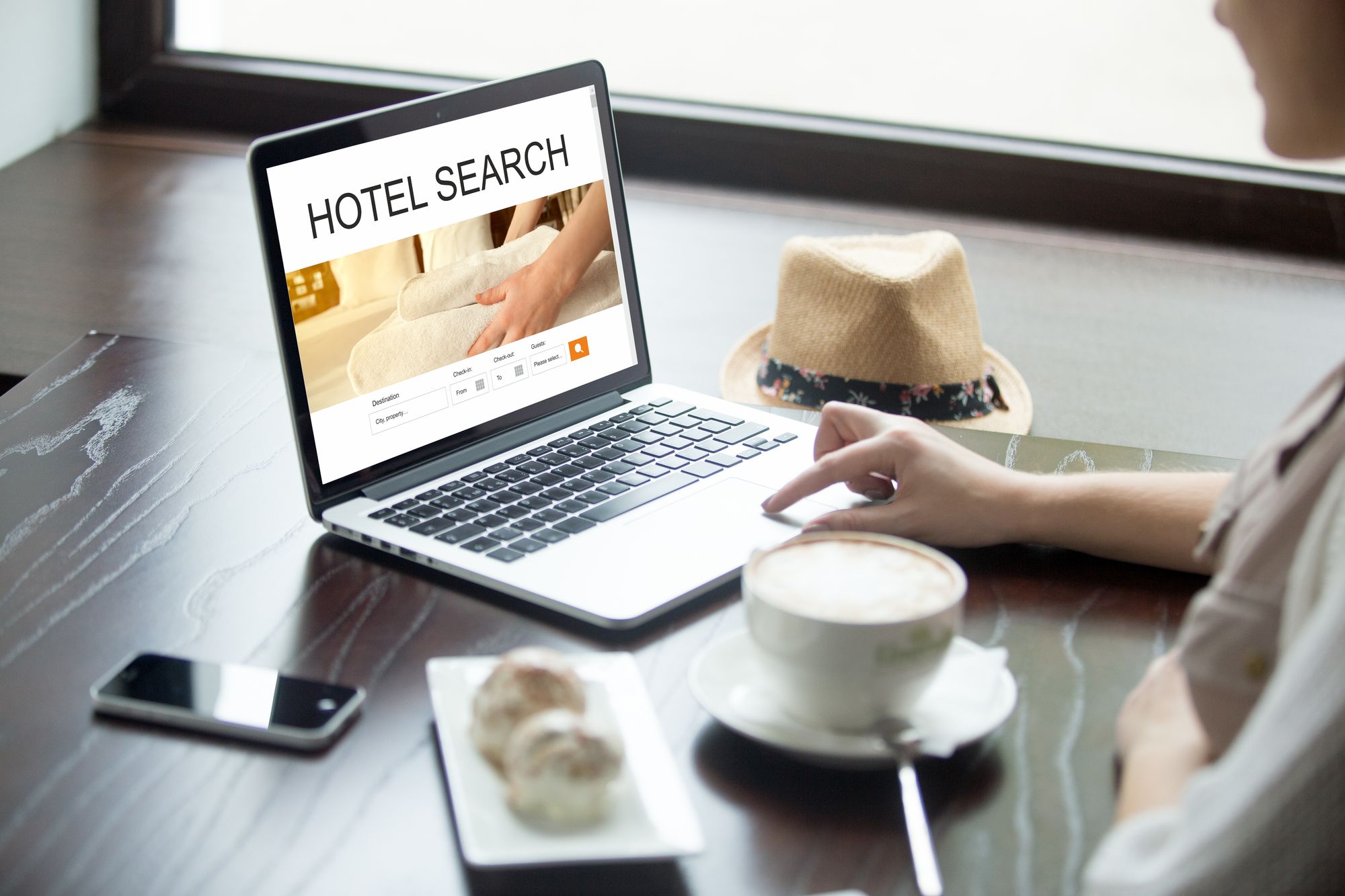 Attractive young woman sitting in cafe with cup of coffee and cakes, working on laptop computer, searching for lodging using on-line web service, booking a hotel on website. View over the shoulder