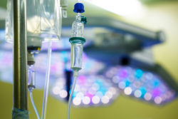 Functioning IV drip hanging on a pole in hospital, with LED surgical lights in the background. Patient, illness, treatment, hospital, medicine and healthcare abstract and concept.