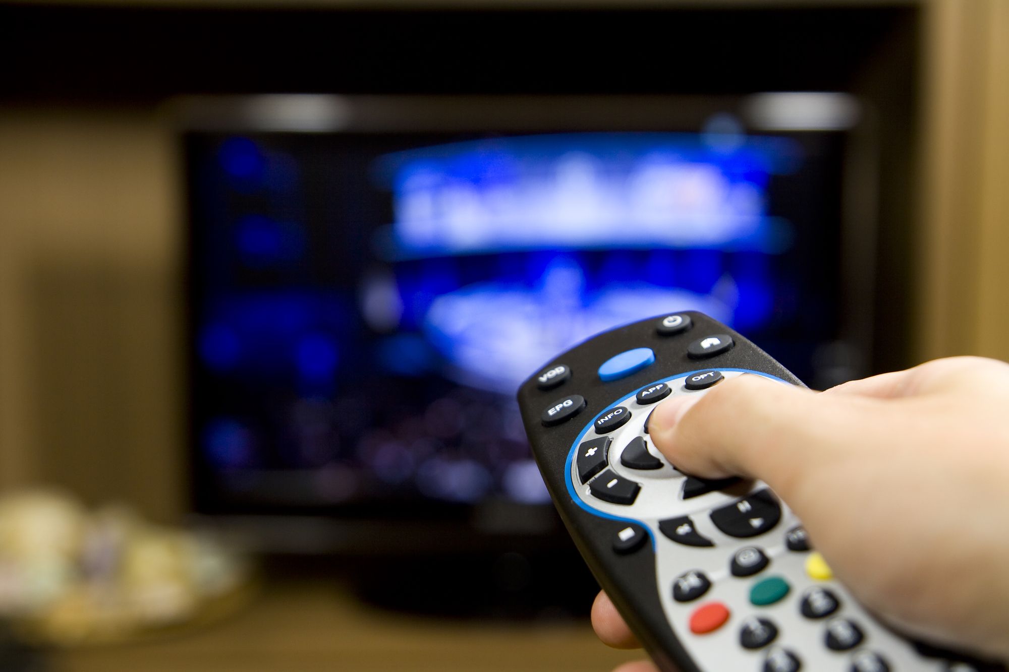 Hand holding TV remote control with a television in the background. Close up.
