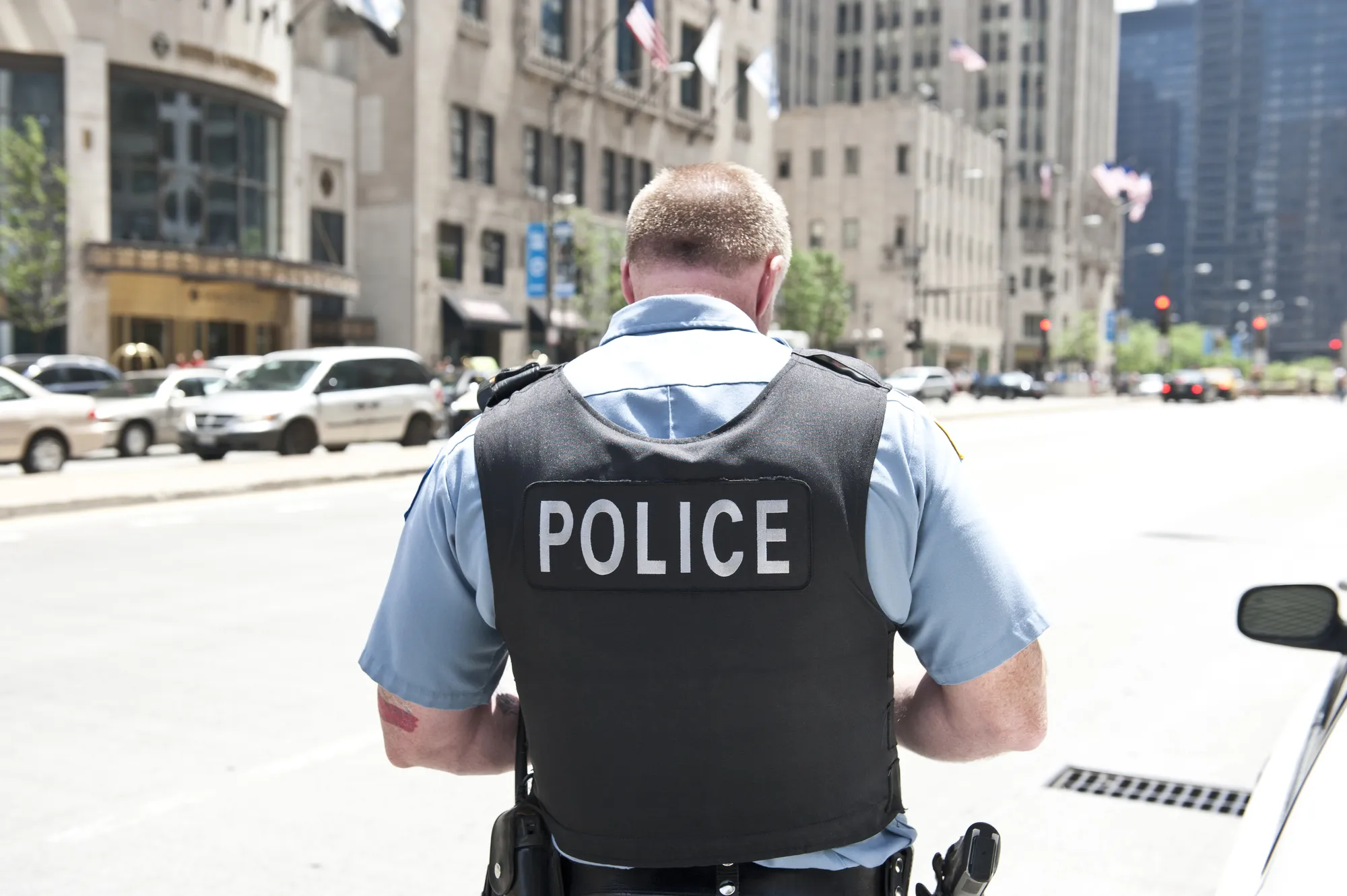 A Chicago city policeman standing on the road on a sunny day wearing a bullet proof vest mentioning Police on his body. On the background, cars and tall buildings are seen.