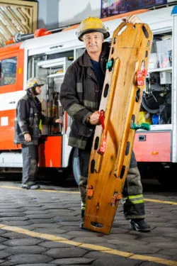 Full length portrait of confident firefighter holding wooden stretcher against truck at fire station