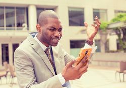 Frustrated handsome young business man in suit receiving bad news message on mobile smart phone standing outdoors outside corporate office
