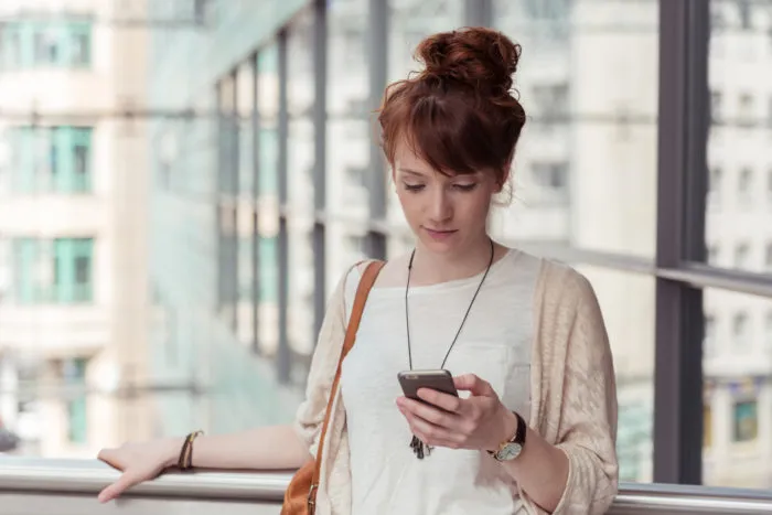 Young woman pausing to check an sms or text message on her mobile phone against a backdrop of high-rise urban buildings