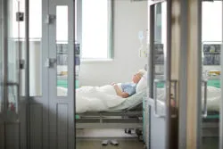 Wide shot image of doors opened to sterile light hospital ward with sad lonely old woman lying on bed in the room