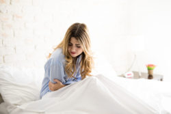 Young Caucasian woman in pajamas grabbing her belly while suffering from stomachache in her bed