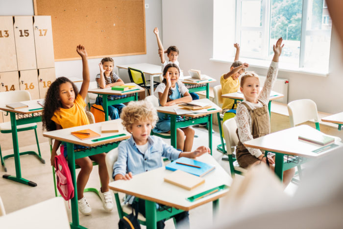 young students in classroom raising their hands to answer a question