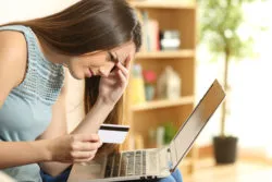 A woman holding a debit card is looking concerned at her laptop.