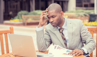 A confused man looks at papers and a laptop.