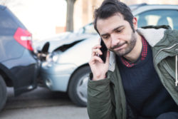 A man is on the phone with two crashed cars behind him.