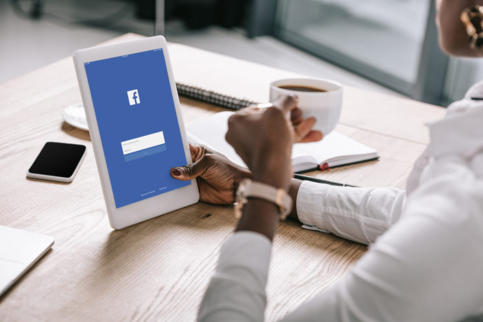 woman holding a cup of coffee in one hand and a tablet in the other. The Facebook app is open on the tablet.