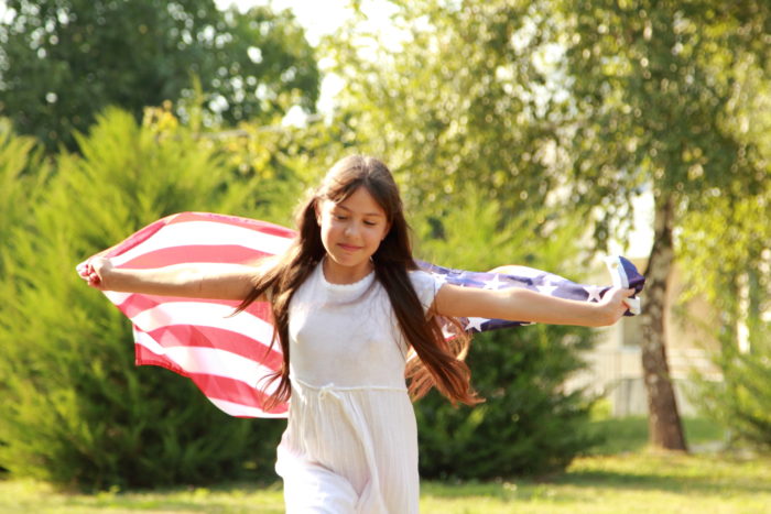 young immigrant girl running through the grass waving an American flag