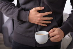 A man holds his stomach with one hand and a cup of coffee with the other.