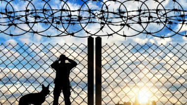 A border guard with a dog looks through a fence.