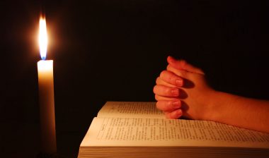 A candle illuminates hands clasped over a bible.