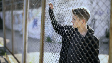 Boy reflects on being detained at CoreCivi Laredo stares through chain link fence.