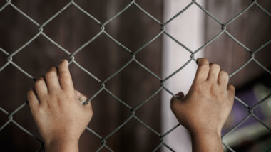 A child's hands hold a chain link fence.