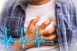 A man in a white t-shirt and blue flannel shirt grabs his chest with his right hand, and a heartbeat graphic overlays the photo - Kombiglyze XR Side Effects