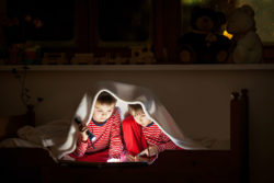 Two boys read by flashlight under a blanket.