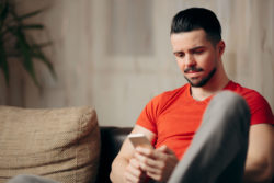 Young man in red t-shirt sits reading texts on smartphone.