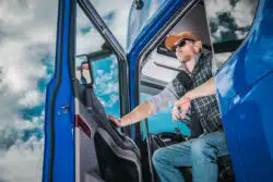 Male trucker in flannel shirt exiting cab of big blue commercial semi truck.