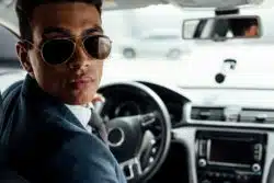 Man in sunglasses looks behind him with his hand on steering wheel as he sits in vehicle's driver's seat