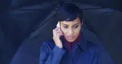 A concerned woman stands in the rain with an umbrella on her cell phone waiting for a ride.