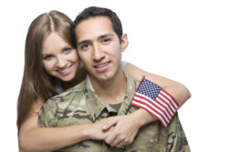 Man in military camo fatigues hugged by wife holding small American flag.