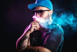 A young man with sunglasses, ear gauges and tattoos shown vaping