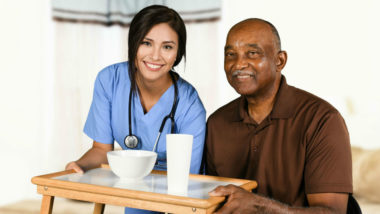 Female health care worker brings food tray to elderly male resident