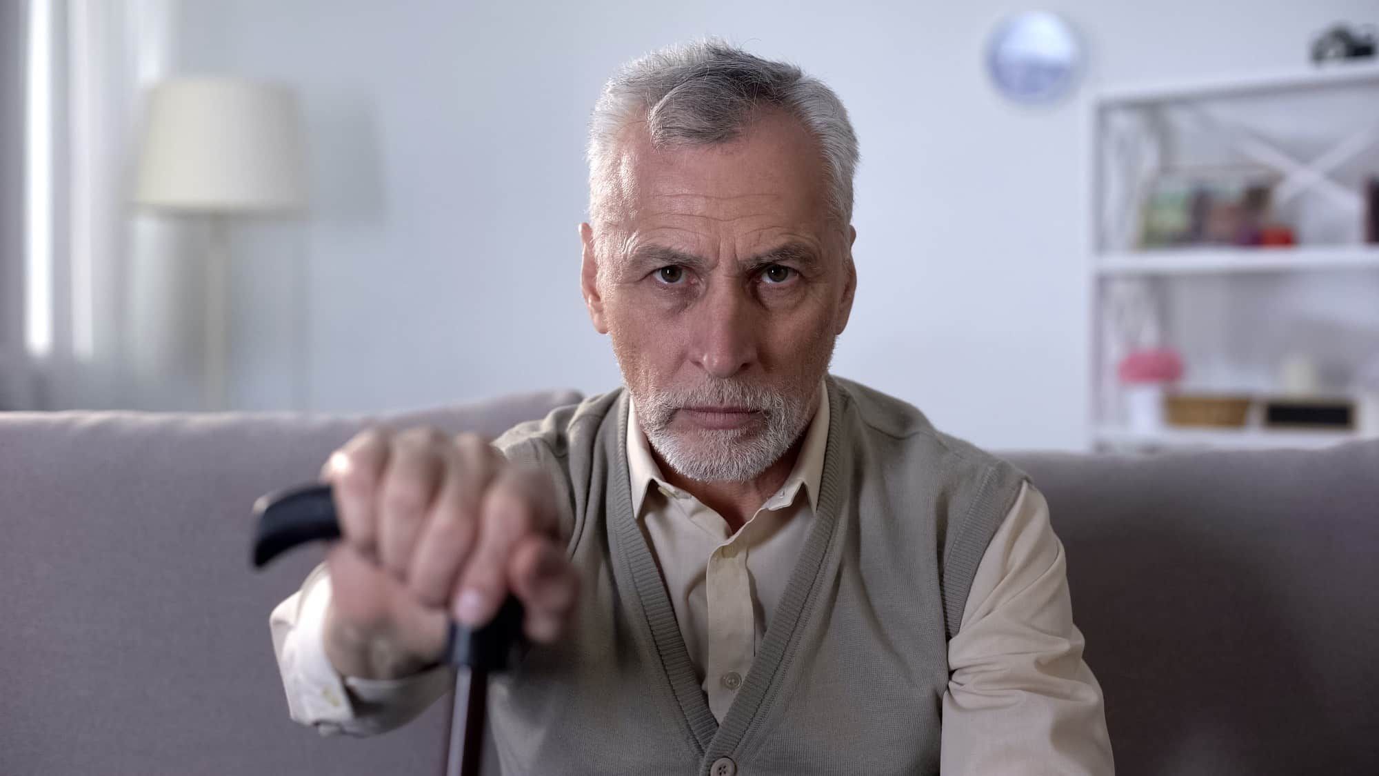 Old man with cane sits on couch and looks at camera