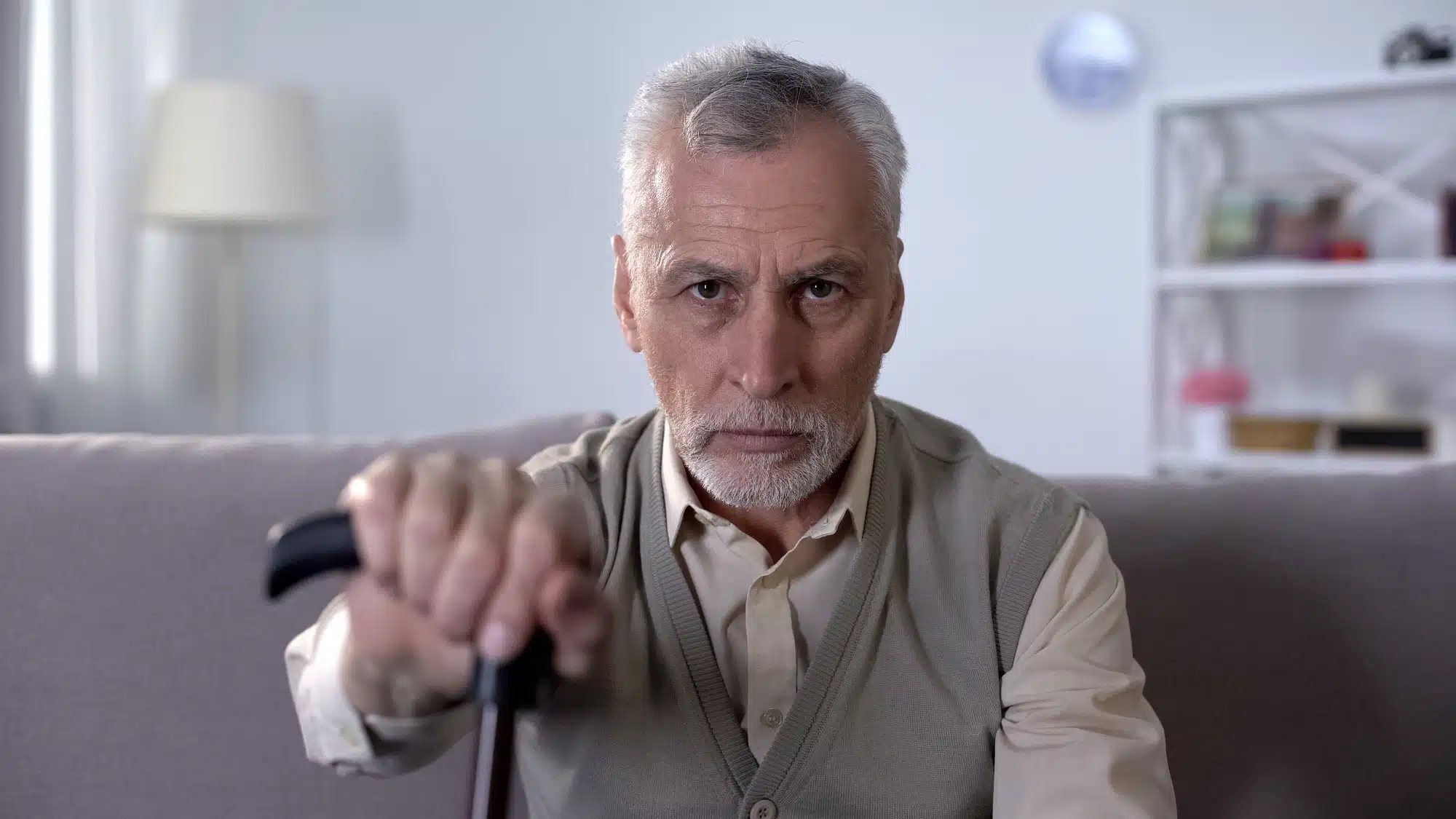 Old man with cane sits on couch and looks at camera