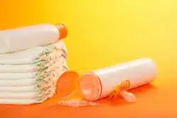 Toppled bottle of baby powder, pacifier and stack of diapers on orange background