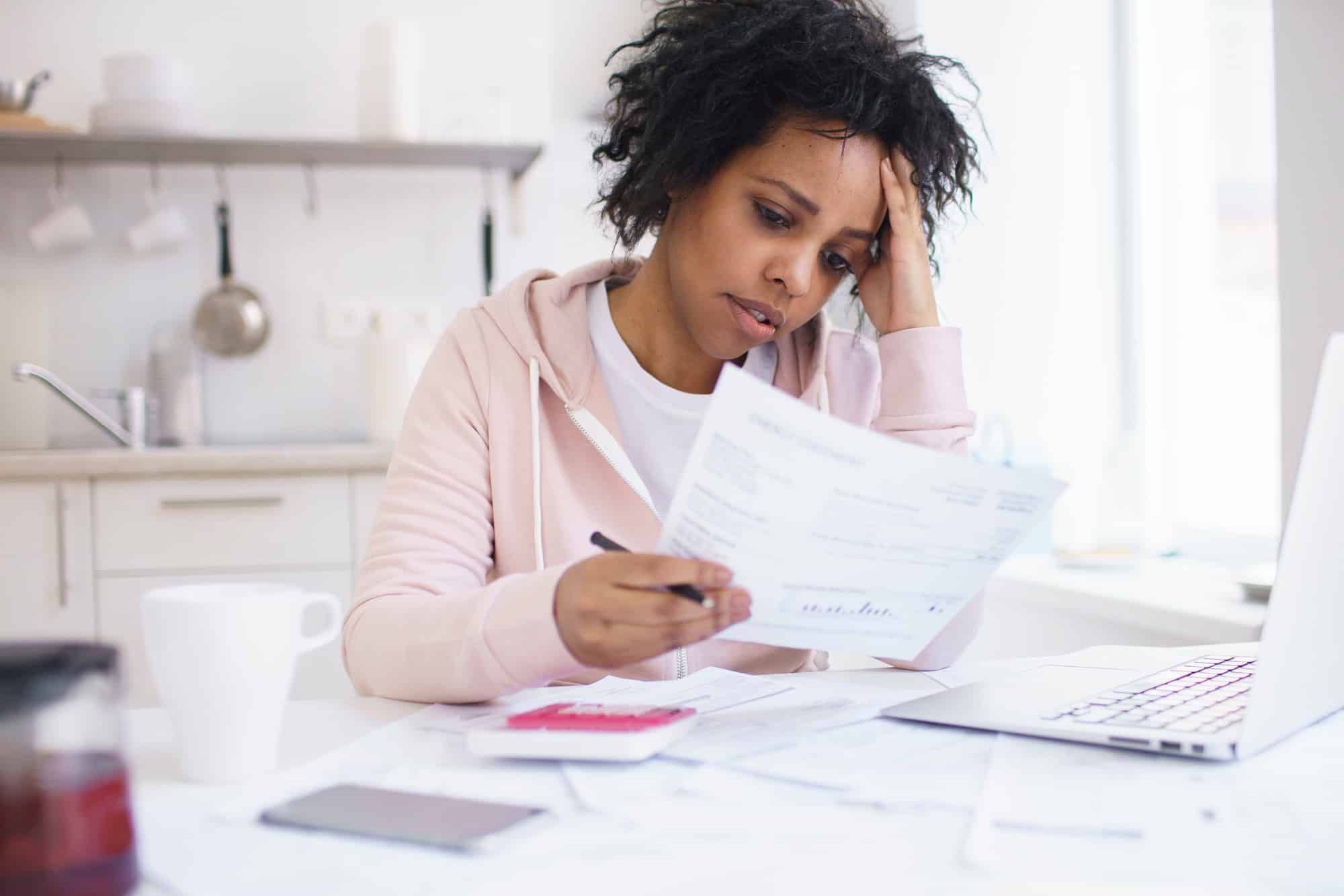 Stressed woman holds invoice at kitchen counter with open laptop