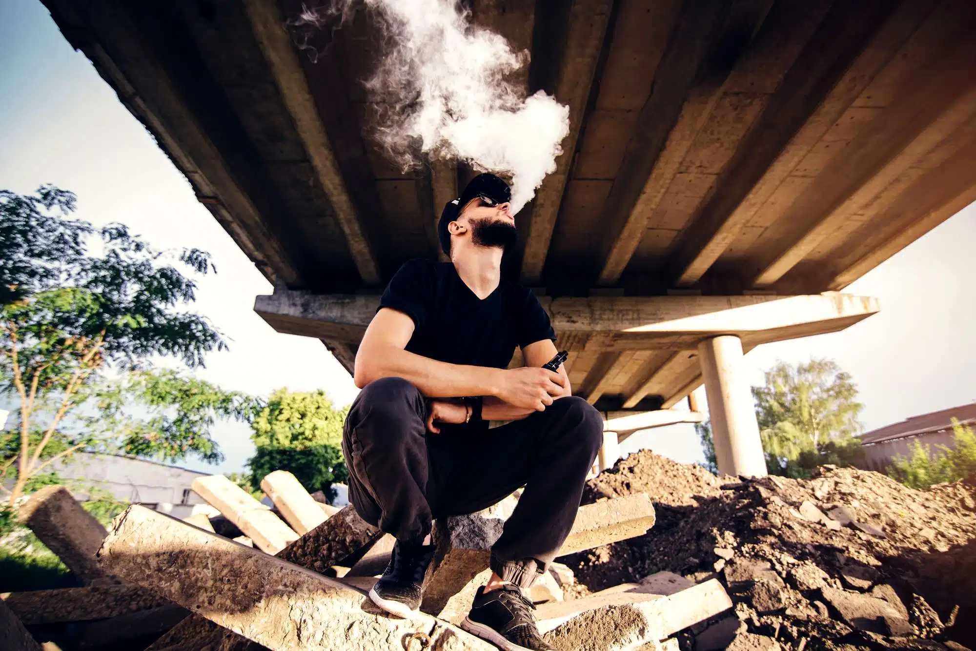 Bearded man sits upon wood pile and vapes