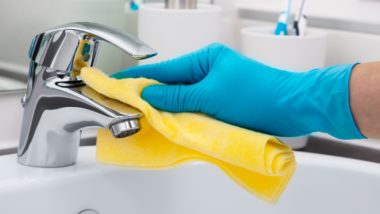 Person wearing a blue rubber glove, cleaning a bathroom sink faucet with a yellow cloth