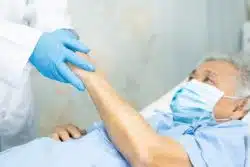 Elderly man in hospital bed and wearing face mask holds gloved doctor's hands