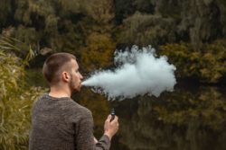 Bearded young man sits near a lake and vapes