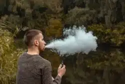 Bearded young man sits near a lake and vapes