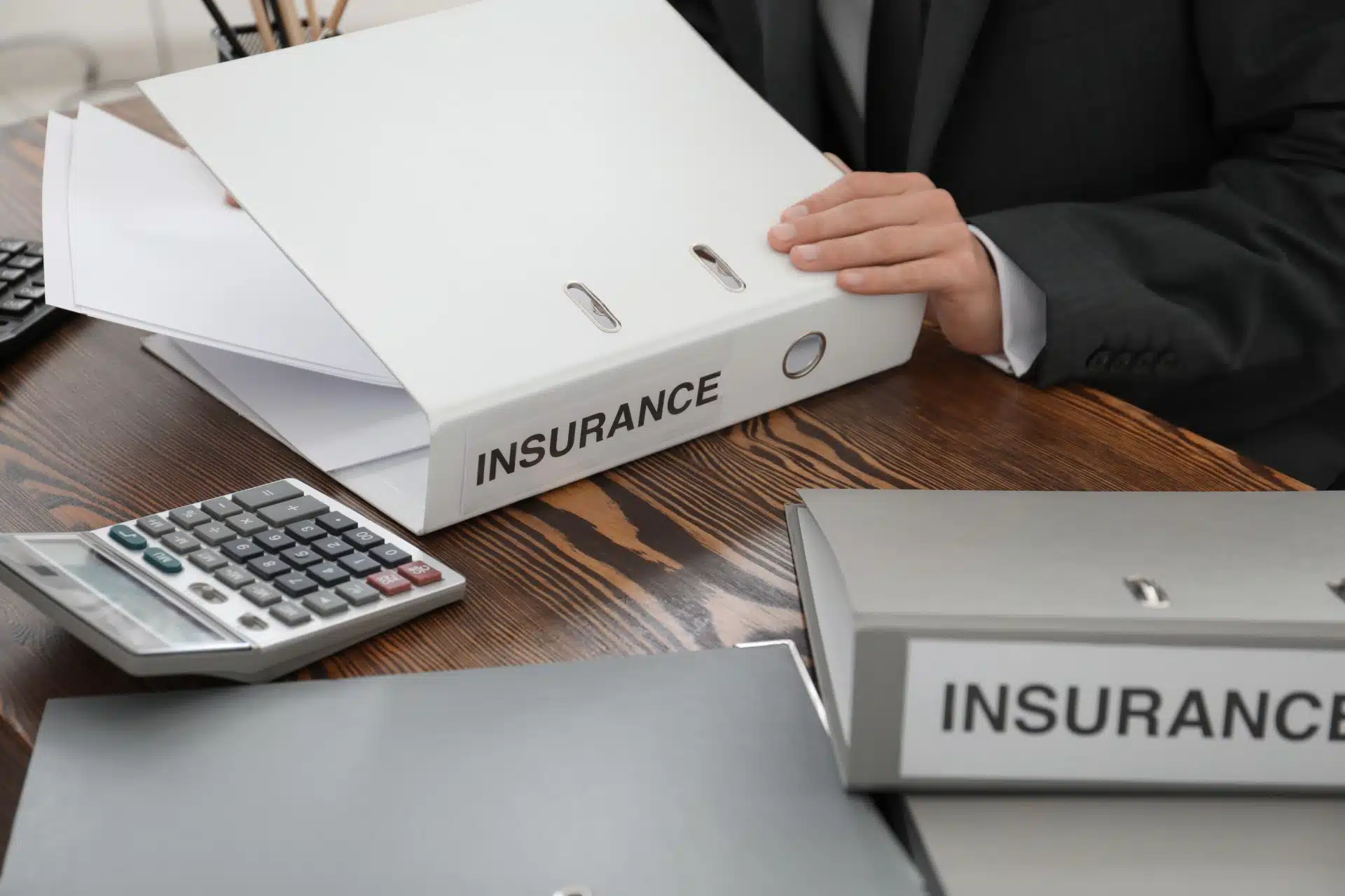 Man in suit opening white "insurance" binder with other binders and calculator on the table