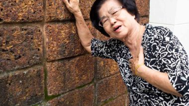 Older woman holding chest with one hand and leaning on wall with other hand