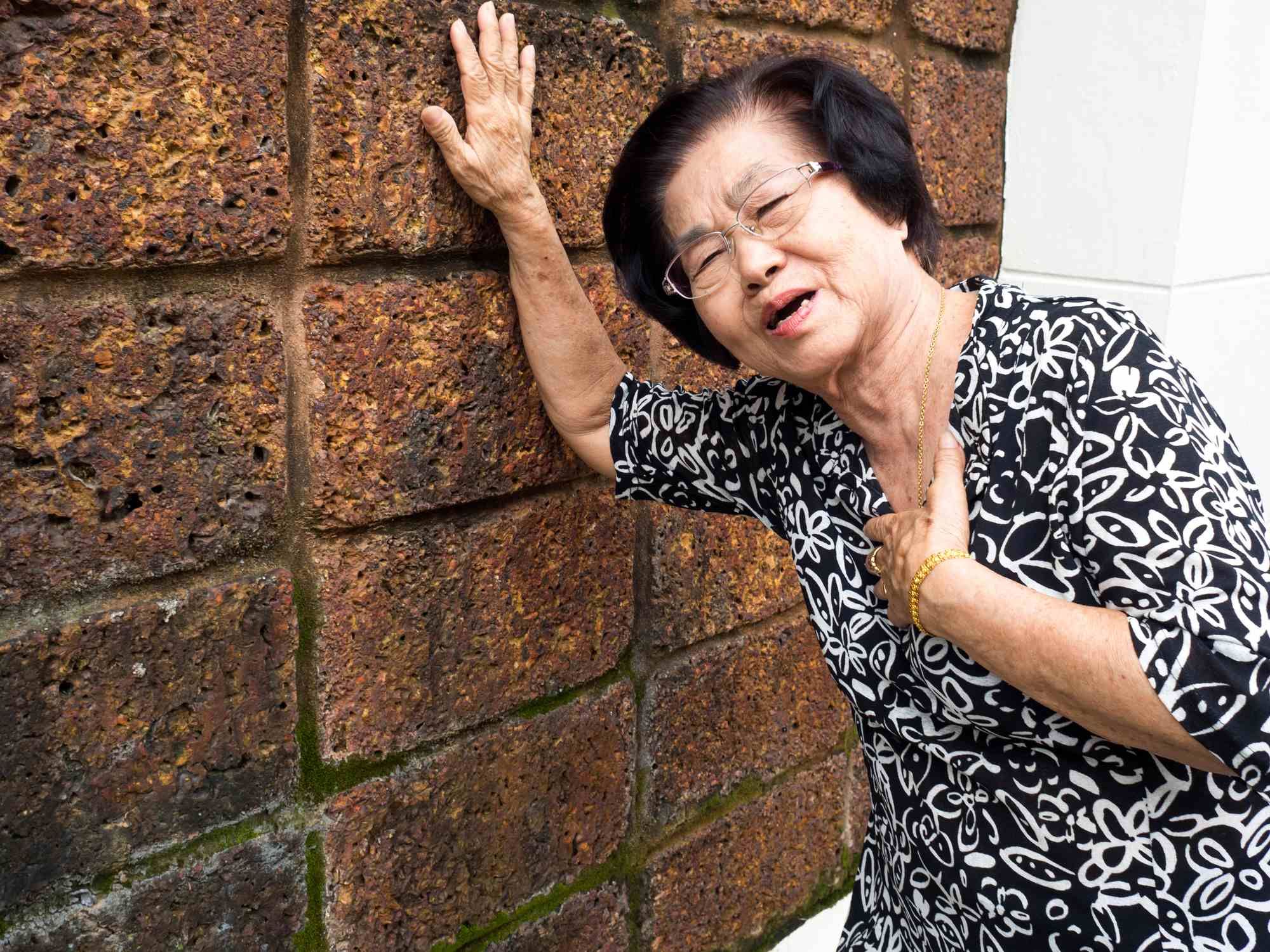 Older woman holding chest with one hand and leaning on wall with other hand