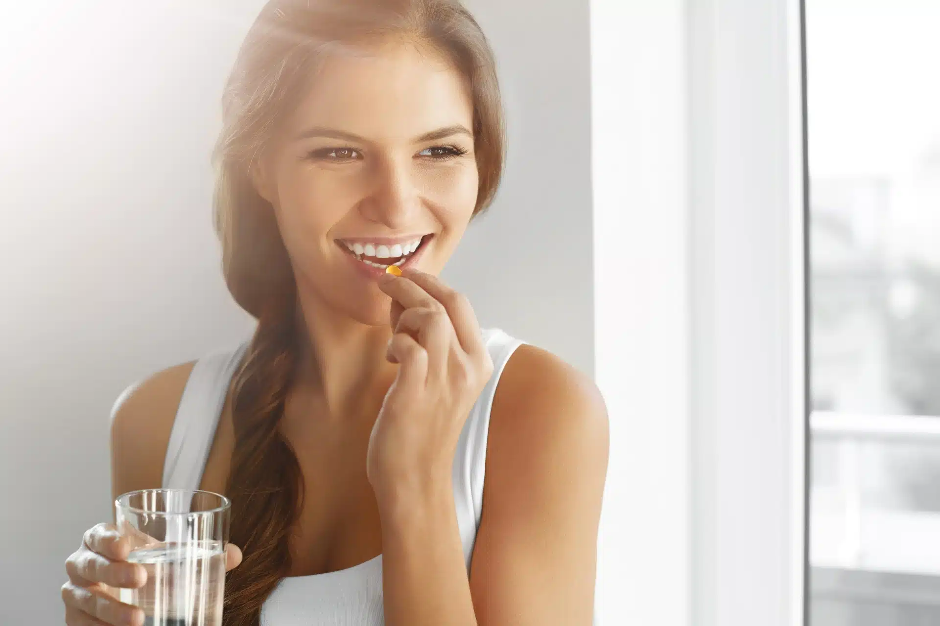 A smiling woman in a white tank top takes a pill while holding a glass of water