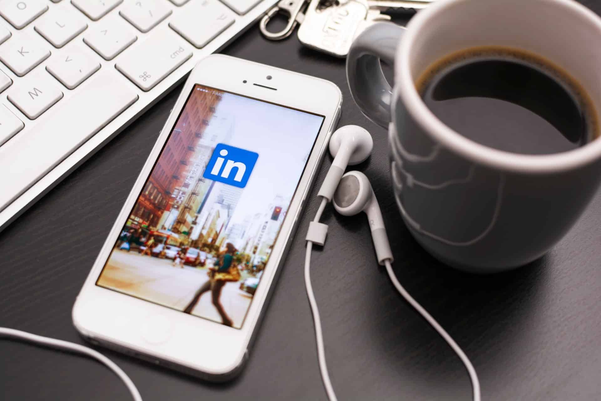 An iPhone displays the LinkedIn app while lying next to coffee mug, keyboard and earbuds