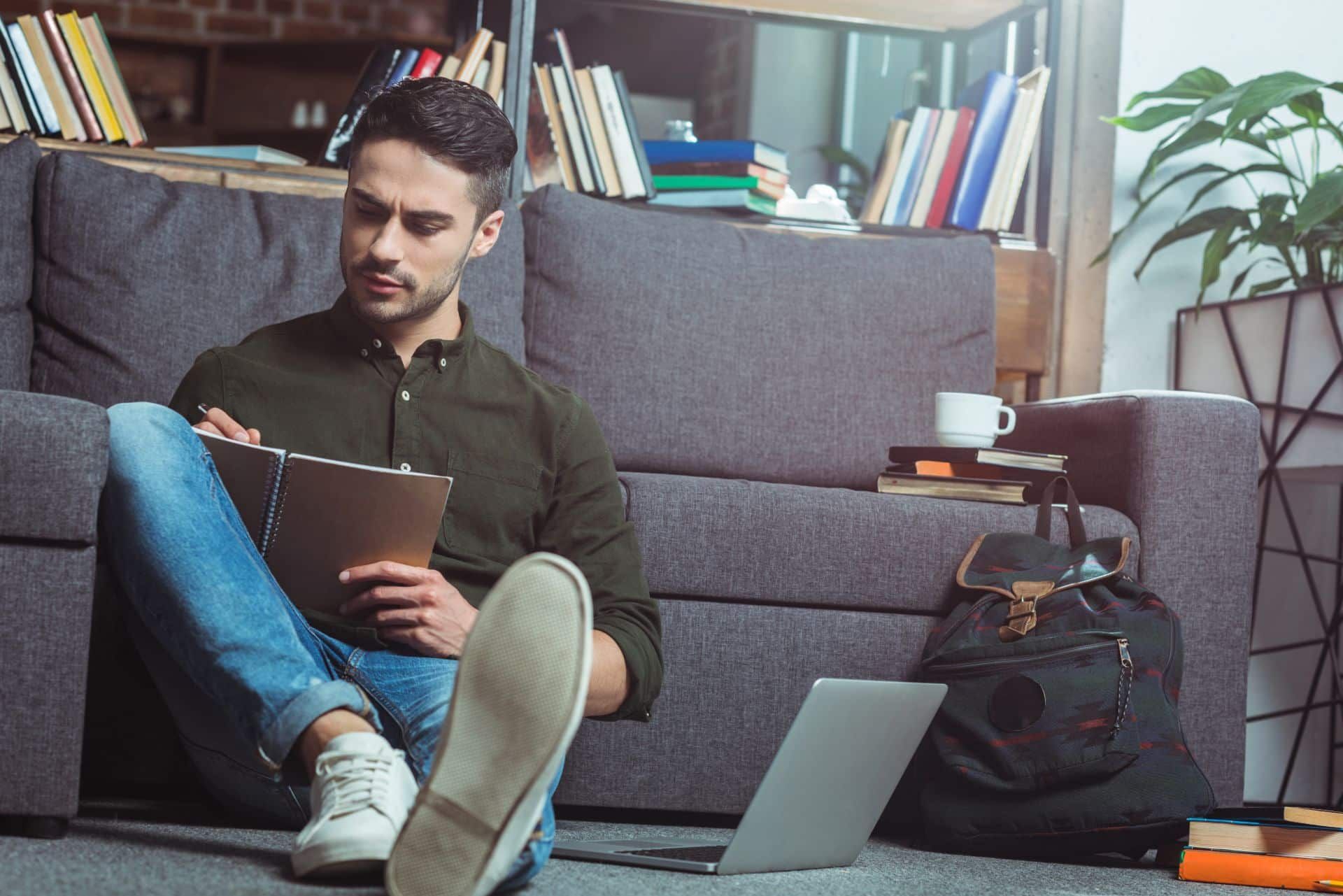 Male adult student sits on floor in library writing notebook, with open laptop, books, coffee cup and backpack