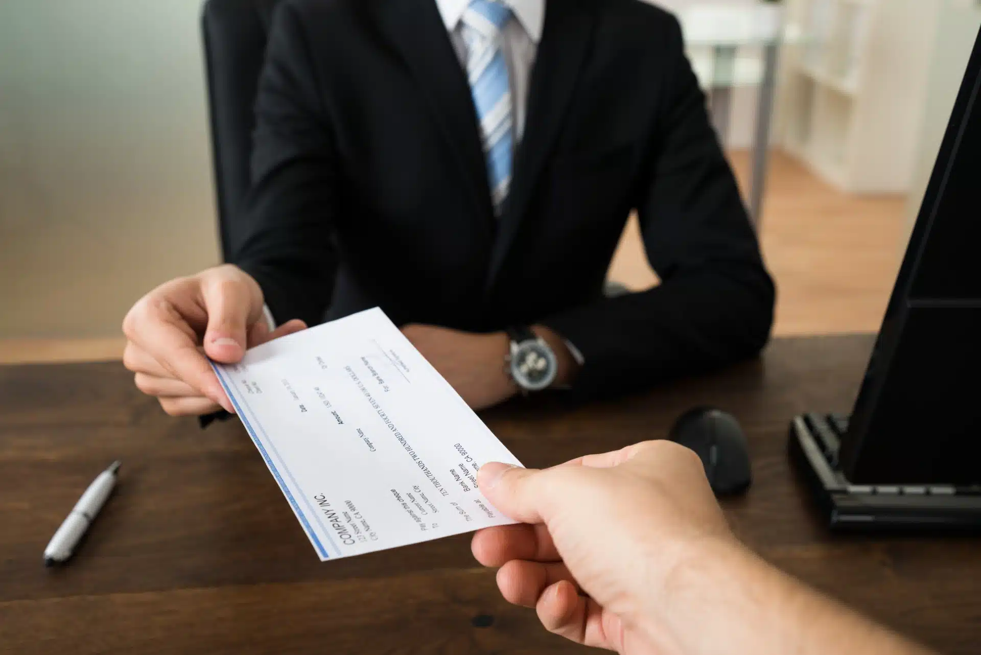 Man in a black suit reaches across a desk to hand someone a check - refund checks