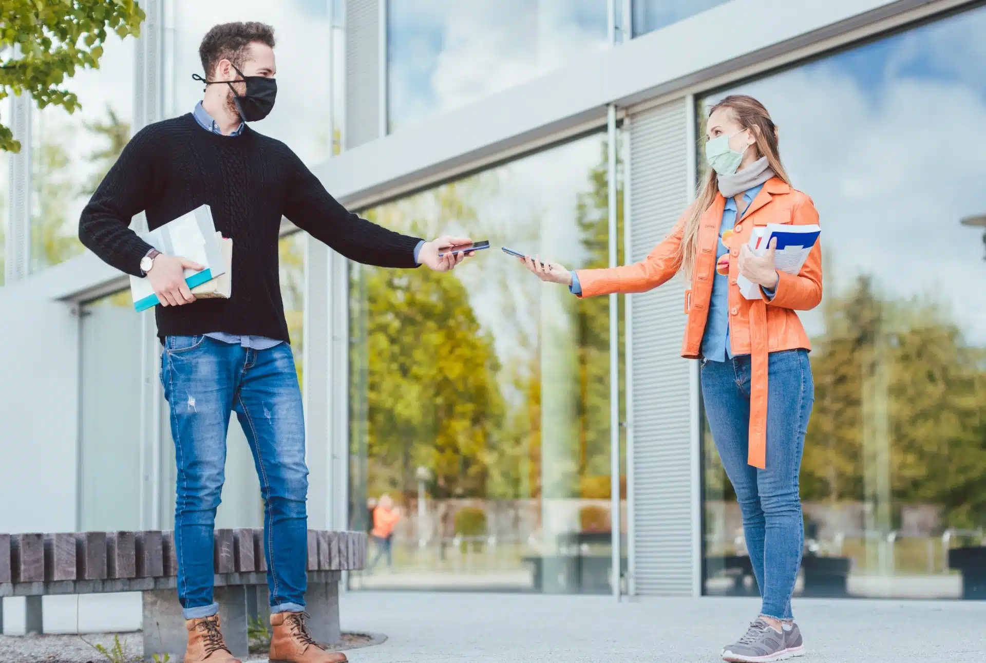 A man and woman holding books outside a building maintain social distancing and hold their smartphones near each other - contact tracing